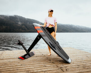 Frau mit E-Foil-Board auf einem Holzsteg vor See und bewaldeten Bergen