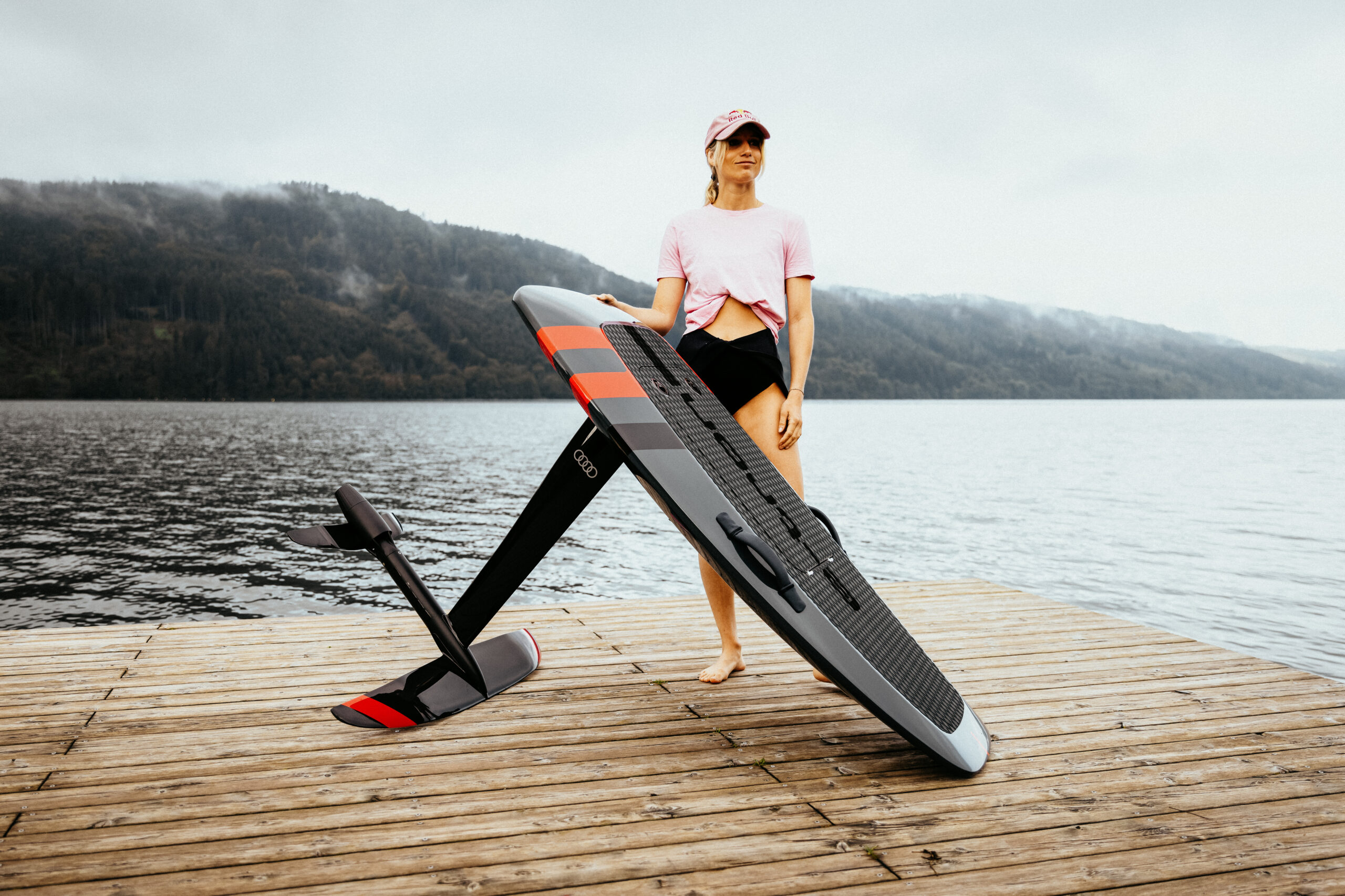 Frau mit E-Foil-Board auf einem Holzsteg vor See und bewaldeten Bergen